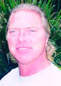A man with light hair and a white shirt smiling in front of green plants.