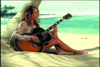 A person sits on a sandy beach playing an acoustic guitar with the ocean in the background.