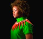 A woman with curly hair wearing a bright green shirt with red pointed collar details against a black background.