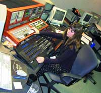 A person sits at a desk adjusting sliders on a large professional audio mixing console surrounded by electronic equipment.