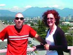 A man in a red shirt and sunglasses and a woman with curly hair smiling in front of a city and mountains.