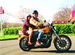 A person in a tan jacket and blue cap drives a motorcycle with a passenger in a dark jacket behind them.