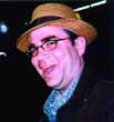 A man with glasses and a straw hat smiling against a dark background.
