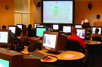 Students sit at desks with computer monitors in a classroom with a large projection screen at the front.