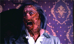 A person with decaying, bloody facial makeup and wide eyes screaming in front of patterned purple wallpaper.