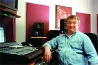A man in a light blue shirt sits in a recording studio next to a mixing console.