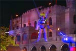 A person in a light-colored costume suspended by wires in front of a large, illuminated castle at night.
