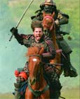 Two people in dark armor riding brown horses toward the camera, one holding a sword aloft.