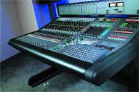 A large professional audio mixing console with numerous sliders, knobs, and buttons on a dark desk.