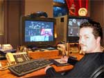 A man sits at a desk facing two computer monitors and a keyboard.
