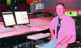 A man in a pink shirt sits beside a large professional audio mixing console and computer monitors.