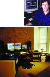 A man smiling in front of a computer and a desk setup with several monitors and a brick wall.