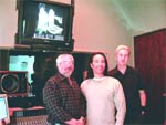 Three people pose in a studio next to a large monitor displaying a graphic and audio equipment.
