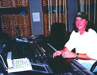 A man in a white shirt and dark cap sits at a large audio mixing desk in a studio.