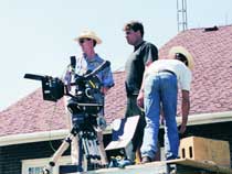 A camera operator, a man standing, and a man in a straw hat working on a film set outdoors.