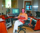A person sits in a chair amidst audio mixing consoles and computer monitors in a studio.