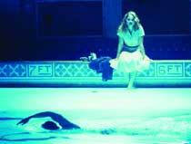 A woman in a light dress sits on a pool edge above a person swimming in blue-tinted water.