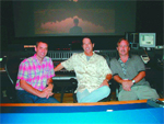 Three men sitting in a studio in front of a large mixing console and a projection screen.