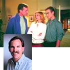 Three people standing together in a room above a portrait of a smiling man with a mustache.