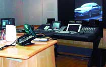 A control room featuring a large mixing console, multiple monitors, and a screen displaying a blue car.