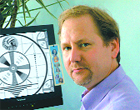 A man in a purple shirt stands beside a monitor showing a black and white circular graphic.