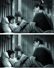 A man and a woman sit together on a bed in a dimly lit room.