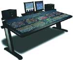 A wide digital audio mixing console with two computer monitors and speakers on a desk.