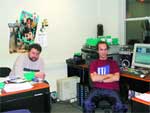 Two men sit in an office with computers, monitors, and posters on the wall.