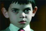 A young boy with dark hair and large ears looks directly at the camera with a serious expression.