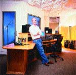 A man in a white shirt and blue jeans leans against a wooden desk in a music studio.