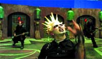 A person with spiked blonde hair and dark makeup performs in a green-lit, laboratory-themed set.