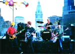 A band performs on an outdoor stage with a tall skyscraper and city buildings in the background.