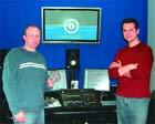 Two men stand in a blue room with a large monitor and audio equipment between them.