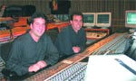 Two men in dark shirts sit behind a large professional audio mixing console in a recording studio.