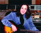 A woman in a blue jacket holding an electric guitar in front of a mixing console.