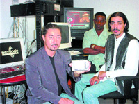 Three men sit in an office with computer monitors and cables in the background.