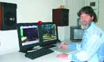 A man with a beard sits at a desk using two computer monitors, a keyboard, and speakers.