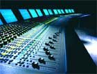 A large professional audio mixing console with numerous knobs, sliders, and glowing blue display screens.