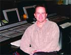 A man in a light-colored shirt sitting in front of a large audio mixing desk and computer monitors.