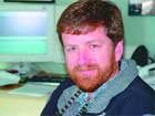 A man with reddish-brown hair and a beard smiling in front of a computer monitor.