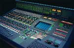 A professional audio mixing console with numerous sliders, knobs, and illuminated buttons.