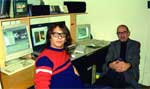 A person in a red sweater and a person in a grey blazer sit in an office with several computer monitors.