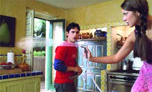 A woman in a pink shirt sprays a man in a red shirt with a water hose in a kitchen.