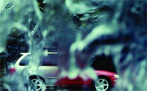 A blurred view of a silver and red car through a window covered in raindrops.