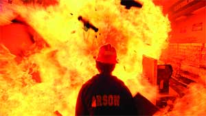 A person in a red hat and black shirt with 'ARSON' text faces a large orange and yellow explosion.