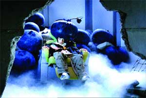 A person lies in a yellow chair surrounded by large blue spheres and thick white fog in a broken concrete room.