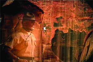 A man in a tan uniform looks toward rock carvings on a cave wall under warm lighting.