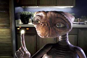 A small, brown, wrinkled alien with large eyes points a glowing finger upward in a dimly lit kitchen.