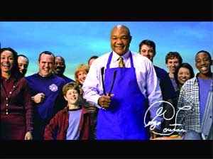 A man in a blue apron and white shirt stands in front of a smiling group of people against a blue sky.