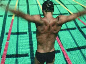 A muscular swimmer in a black cap and trunks stands in a pool with arms spread wide.